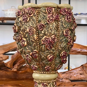 Op de afbeelding: Een decoratieve keramische vaas met een gegolfde rand en een voet. De vaas is bedekt met een reliëfdesign van rozen en bladeren in tinten groen, goud en bordeaux.