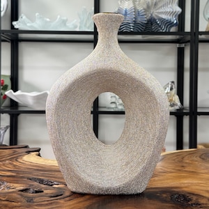 Könnte beinhalten: Eine strukturierte, beige Keramikvase mit einer großen ovalen Öffnung in der Mitte. Die Vase hat einen schmalen Hals und einen breiten, abgerundeten Körper. Die Vase steht auf einer Holzoberfläche.
