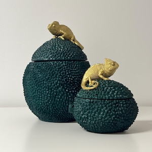 May include: Two dark teal decorative jars with textured surfaces, each topped with a gold-coloured chameleon figurine. The jars are different sizes and have lids. Plain white background.