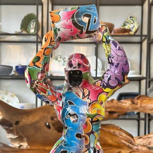 Peut inclure: Une sculpture de gorille colorée de style graffiti avec un bras levé tenant un objet carré. La sculpture est en résine et est peinte dans une variété de couleurs vives, notamment le rouge, le bleu, le vert, le jaune et le violet.