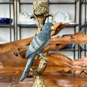 May include: A gold candle holder with a blue and white bird perched on a branch. The bird has a gold beak and is facing to the right. The candle holder has a gold base and a gold candle cup.