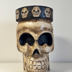 Puede incluir: Una gran escultura de calavera tallada con un acabado blanquecino envejecido. La calavera tiene cuencas de ojos grandes y una mandíbula detallada. Una banda negra con detalles de calaveras rodea la parte superior. Esta pieza decorativa es ideal para Halloween o decoración gótica.