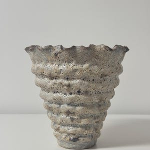 May include: A textured ceramic vase with a flared, ruffled rim. The vase has a light gray and brown color scheme with a bubbly, porous surface. The body of the vase is a series of stacked, rounded ridges.