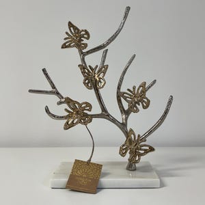May include: A decorative metal sculpture featuring a tree branch design with gold-colored butterflies. The sculpture is mounted on a white marble base. A small tag is attached to the sculpture.