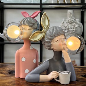 May include: Two decorative figurines of women blowing bubbles with illuminated orbs. One wears a pink dress with white polka dots and a pink bow. The other wears a grey turtleneck and holds a coffee cup. Shelves in the background display decorative items.