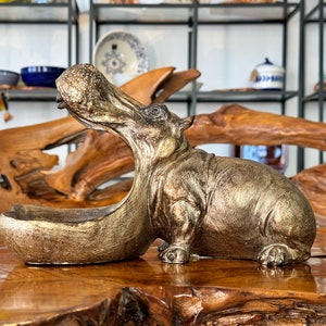 Könnte beinhalten: Eine goldfarbene Flusspferd-Figur mit offenem Maul, die als dekorative Schale dient. Die Figur steht auf einer Holzoberfläche.