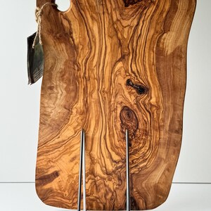 May include: A large, irregularly shaped wooden cutting board with a natural wood grain pattern. The board has a hole in the upper left corner and is displayed on a metal stand. The wood tones range from light to dark brown.