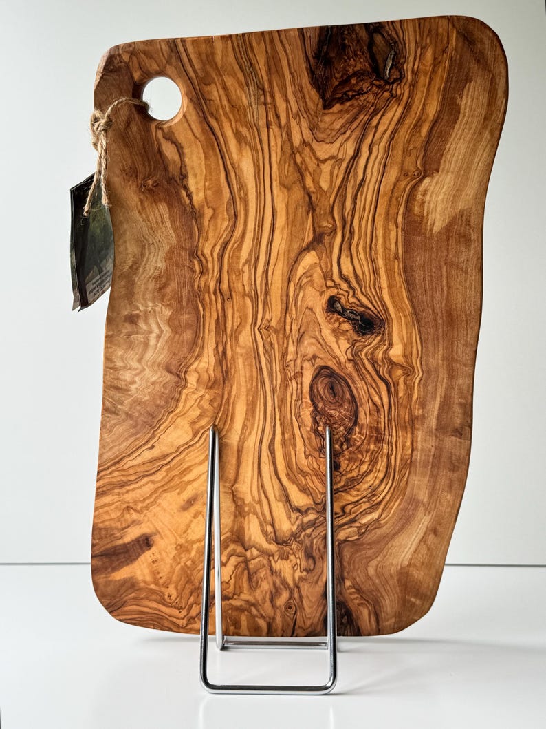 May include: A large, irregularly shaped wooden cutting board with a natural wood grain pattern. The board has a hole in the upper left corner and is displayed on a metal stand. The wood tones range from light to dark brown.