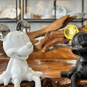 Puede incluir: Dos figuras de Mickey Mouse. Una es blanca con orejas plateadas, la otra es negra con orejas doradas. Ambas est&aacute;n sentadas sobre una superficie de madera. El fondo incluye estantes con platos decorativos.