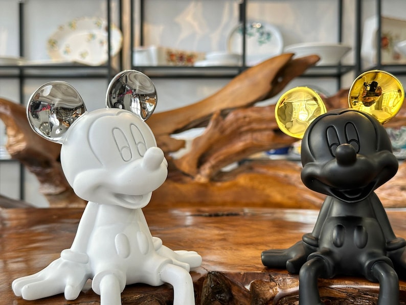 Puede incluir: Dos figuras de Mickey Mouse. Una es blanca con orejas plateadas, la otra es negra con orejas doradas. Ambas est&aacute;n sentadas sobre una superficie de madera. El fondo incluye estantes con platos decorativos.