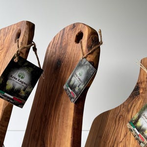 May include: Three wooden cutting boards with natural wood grain and a hole for hanging. Each board has a tag with the text "Arte Legno" and the Italian flag. The boards are made in Italy.