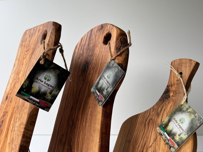 May include: Three wooden cutting boards with natural wood grain and a hole for hanging. Each board has a tag with the text "Arte Legno" and the Italian flag. The boards are made in Italy.
