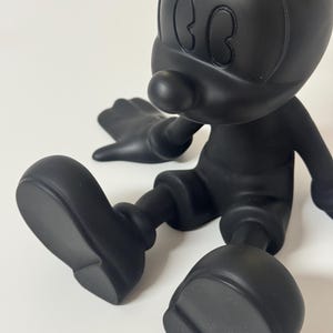 Pu&ograve; includere: Statuetta nera opaca di un personaggio dei cartoni animati con grandi orecchie, naso rotondo e pantaloncini. La figura &egrave; seduta con le gambe distese e le braccia lungo i fianchi. Una piccola porzione di una corona color oro &egrave; visibile sulla testa.