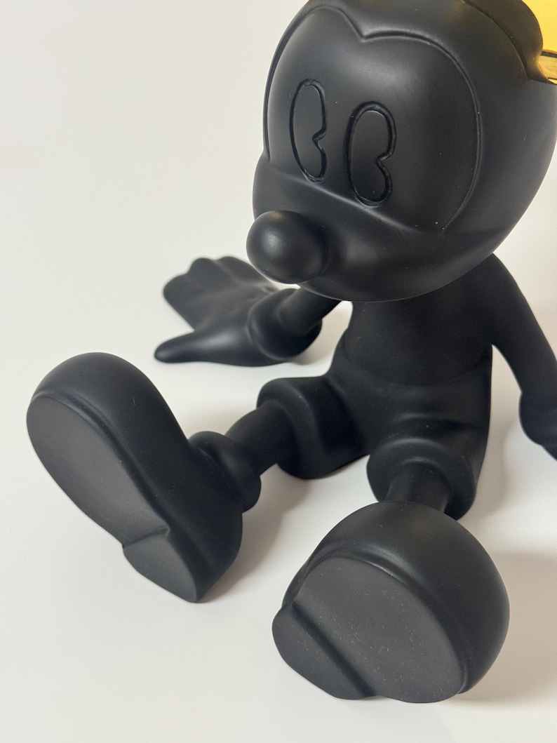 Pu&ograve; includere: Statuetta nera opaca di un personaggio dei cartoni animati con grandi orecchie, naso rotondo e pantaloncini. La figura &egrave; seduta con le gambe distese e le braccia lungo i fianchi. Una piccola porzione di una corona color oro &egrave; visibile sulla testa.