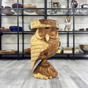 May include: A wooden owl stool with a carved design. The owl has a brown and beige color scheme and is sitting on a wooden base.