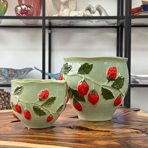 Può includere: Due vasi in ceramica verde con disegni di fragole in rilievo. Il vaso più grande è più alto e più largo del vaso più piccolo. Entrambi i vasi hanno una smaltatura verde chiaro e presentano fragole rosse con foglie verdi.