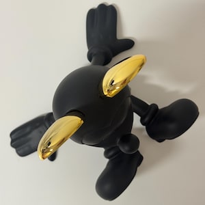 Pu&ograve; includere: Una statuetta nera con testa, braccia e gambe arrotondate. La statuetta ha accenti color oro sulla testa ed &egrave; posizionata con le braccia e le gambe tese. La figura &egrave; ambientata su uno sfondo bianco.