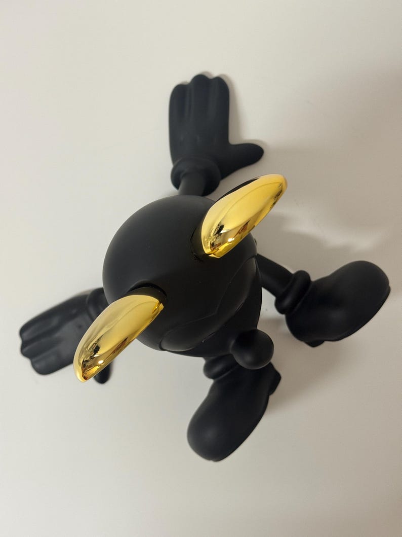 Pu&ograve; includere: Una statuetta nera con testa, braccia e gambe arrotondate. La statuetta ha accenti color oro sulla testa ed &egrave; posizionata con le braccia e le gambe tese. La figura &egrave; ambientata su uno sfondo bianco.