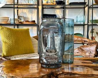 May include: Two glass vases, one with a face design and the other clear, on a wooden table. The vase with the face design is blue and gray, while the clear vase is light blue.