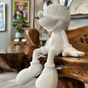 Puede incluir: Una figura blanca de Mickey Mouse con orejas plateadas brillantes sentada sobre una superficie de madera. La figura tiene un dise&ntilde;o cl&aacute;sico, con guantes blancos grandes, pantalones cortos y zapatos. El fondo incluye obras de arte y estanter&iacute;as.