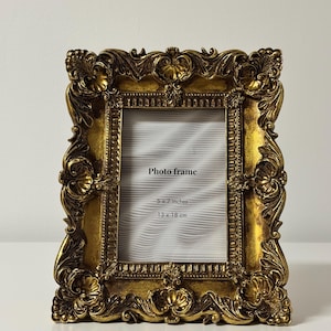 Pode incluir: Moldura dourada ornamentada com abertura retangular. A moldura apresenta um design esculpido detalhado com motivos florais e de rolagem. A moldura mede 13 x 18 cm e tem as palavras "Photo frame" impressas no interior.