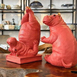 Könnte beinhalten: Drei rote Nilpferdfiguren in Yoga-Posen. Eines ist kopfüber, ein anderes sitzt und das dritte liegt auf einem Buch. Die Flusspferde haben goldene Akzente an Ohren und Füßen. Die Figuren stehen auf einem Holztisch.