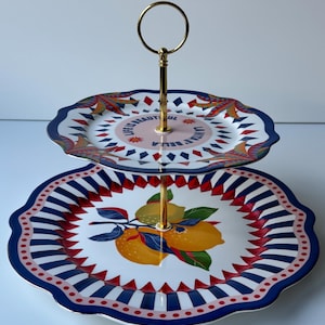 Könnte beinhalten: Zweistöckige Servierplatte mit goldenem Griff. Die obere Ebene hat einen weißen Hintergrund mit einem farbenfrohen Design und dem Text "LIFE IS BEAUTIFUL LA VITA BELLA". Die untere Ebene hat ein Zitronenmuster mit blauen, roten und weißen Streifen.