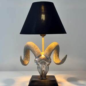 May include: A decorative table lamp featuring a ram skull design. The lamp has a black lampshade and a silver-colored ram skull base. The base is square and dark-colored. The lamp is lit, casting a warm glow.