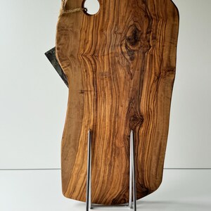 May include: A wooden cutting board with a natural edge, featuring a hole for hanging and a dark-colored knife. The board is displayed on a silver metal stand. The wood grain is prominent, showcasing a warm, brown color.