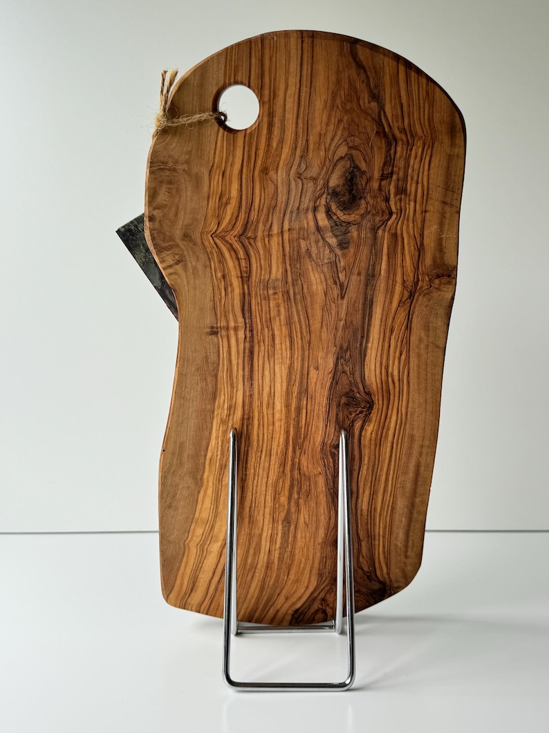 May include: A wooden cutting board with a natural edge, featuring a hole for hanging and a dark-colored knife. The board is displayed on a silver metal stand. The wood grain is prominent, showcasing a warm, brown color.
