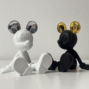 Pu&ograve; includere: Due figurine stilizzate di Topolino. Una &egrave; bianca con orecchie argentate riflettenti, l'altra nera con orecchie dorate. Entrambe sono sedute con le gambe distese, le braccia lungo i fianchi e un classico volto di Topolino.