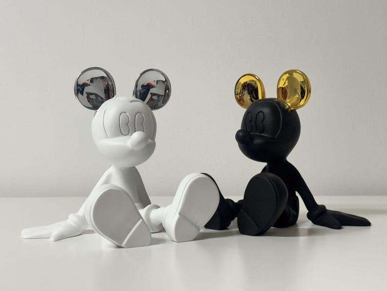 Pu&ograve; includere: Due figurine stilizzate di Topolino. Una &egrave; bianca con orecchie argentate riflettenti, l'altra nera con orecchie dorate. Entrambe sono sedute con le gambe distese, le braccia lungo i fianchi e un classico volto di Topolino.