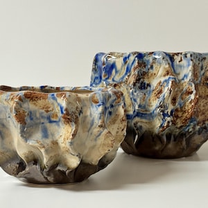 Könnte beinhalten: Zwei Keramikschalen mit einem strukturierten, organischen Design. Die Schalen weisen eine Mischung aus Farben auf, darunter Braun, Blau und Creme, mit einem glänzenden Finish. Die Schalen haben eine einzigartige, unregelmäßige Form, die auf handwerkliche Herkunft hindeutet.