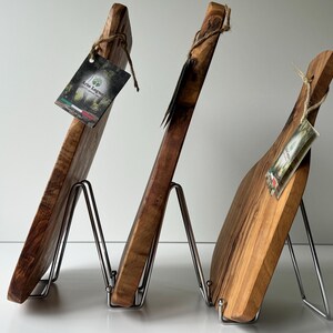 May include: Three wooden cutting boards of varying shapes and sizes, displayed on metal stands. The boards are made of light and dark brown wood, with visible grain patterns. Each board has a tag attached with twine.