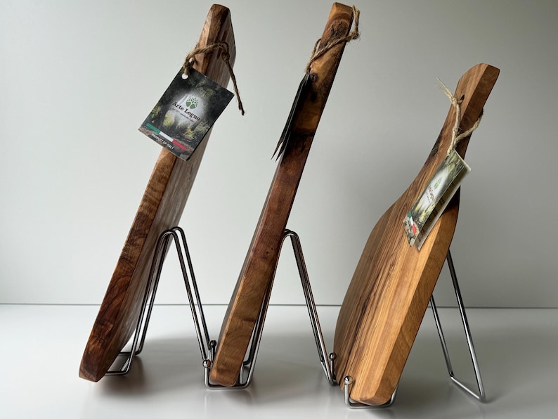 May include: Three wooden cutting boards of varying shapes and sizes, displayed on metal stands. The boards are made of light and dark brown wood, with visible grain patterns. Each board has a tag attached with twine.