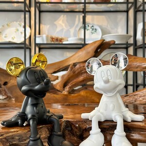 Puede incluir: Dos figuras de Mickey Mouse, una negra y otra blanca, est&aacute;n una al lado de la otra. La figura negra tiene orejas doradas, mientras que la blanca tiene orejas plateadas. Ambas est&aacute;n sentadas con las piernas colgando, sobre una superficie de madera.