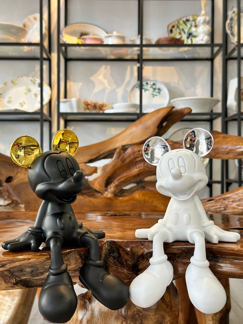 Puede incluir: Dos figuras de Mickey Mouse, una negra y otra blanca, est&aacute;n una al lado de la otra. La figura negra tiene orejas doradas, mientras que la blanca tiene orejas plateadas. Ambas est&aacute;n sentadas con las piernas colgando, sobre una superficie de madera.