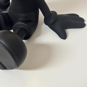Pu&ograve; includere: Una figura nera stilizzata con arti arrotondati e una mano guantata. La figura &egrave; in posa seduta, con una mano appoggiata su una superficie bianca. Il design della figura &egrave; liscio e opaco.