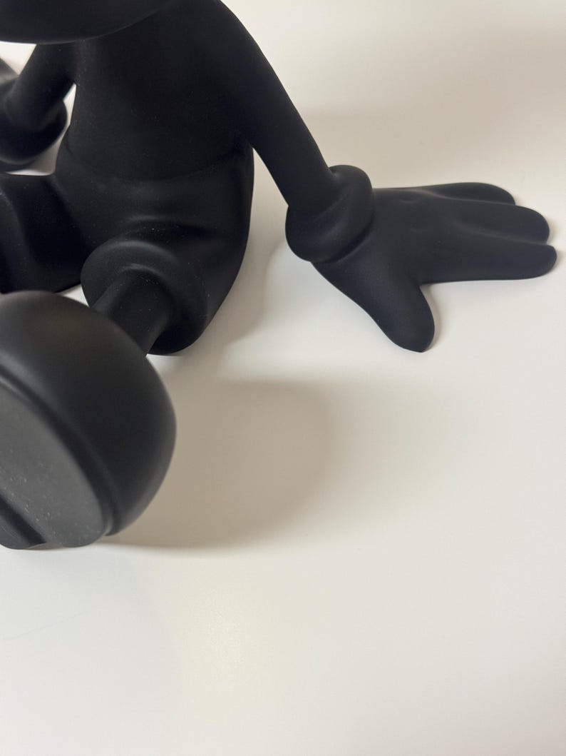 Pu&ograve; includere: Una figura nera stilizzata con arti arrotondati e una mano guantata. La figura &egrave; in posa seduta, con una mano appoggiata su una superficie bianca. Il design della figura &egrave; liscio e opaco.
