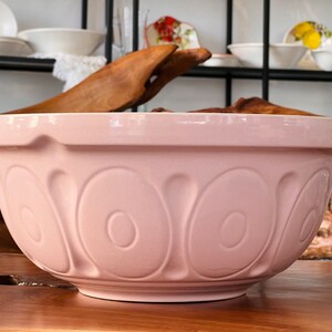 May include: A large, pink ceramic mixing bowl with a decorative, embossed pattern. The bowl is sitting on a wooden surface.