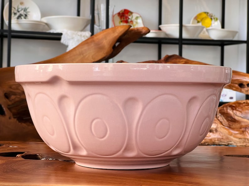 May include: A large, pink ceramic mixing bowl with a decorative, embossed pattern. The bowl is sitting on a wooden surface.