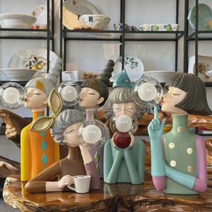 Peut inclure: Ensemble de figurines colorées et fantaisistes représentant des femmes soufflant des bulles. Chaque figurine a une tenue et une coiffure uniques, les bulles contenant une source de lumière. Les figurines sont exposées sur une table en bois.
