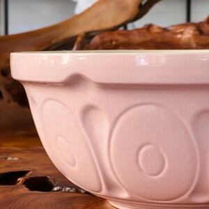May include: A large, pale pink ceramic mixing bowl with a fluted rim and a decorative pattern of raised ovals. The bowl is sitting on a wooden surface.