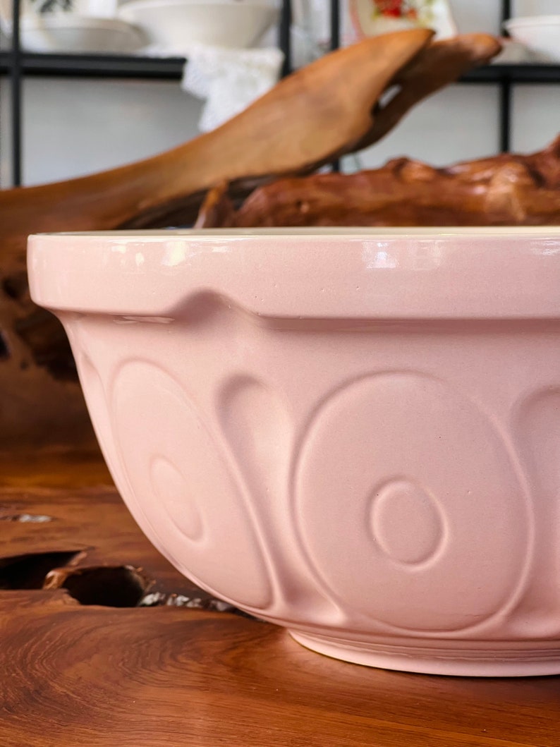 May include: A large, pale pink ceramic mixing bowl with a fluted rim and a decorative pattern of raised ovals. The bowl is sitting on a wooden surface.