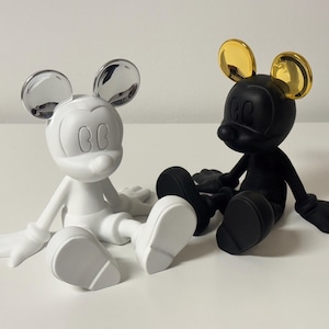 Pu&ograve; includere: Due figurine stilizzate di Topolino. Una &egrave; bianca con orecchie argentate, l'altra &egrave; nera con orecchie dorate. Entrambe sono sedute con le gambe distese. Su una superficie bianca.