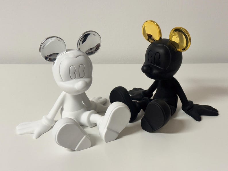 Pu&ograve; includere: Due figurine stilizzate di Topolino. Una &egrave; bianca con orecchie argentate, l'altra &egrave; nera con orecchie dorate. Entrambe sono sedute con le gambe distese. Su una superficie bianca.