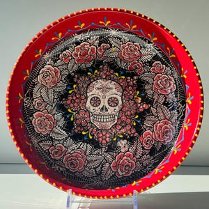 Può includere: Un vassoio rotondo in metallo rosso con un design a teschio di zucchero del Giorno dei Morti. Il teschio è circondato da rose rosa e accenti floreali su sfondo nero. Il bordo del vassoio presenta un motivo decorativo giallo e blu.