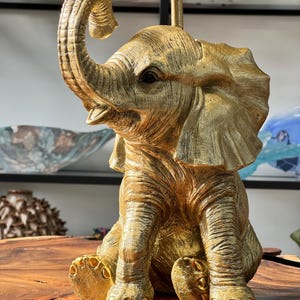 May include: A gold-painted elephant figurine with a raised trunk, sitting on a wooden surface. The elephant has a detailed textured surface and is a decorative accent.