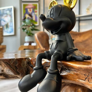 Puede incluir: Una figura negra de Mickey Mouse con orejas doradas, sentada sobre una superficie de madera. La figura tiene un acabado mate y est&aacute; posada con las piernas colgando. El fondo incluye obras de arte y un vistazo a un interior moderno.