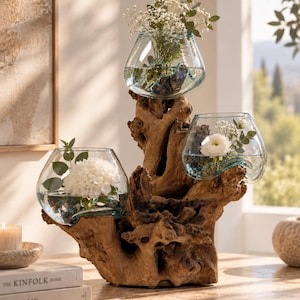 Könnte beinhalten: Ein dekoratives Herzstück bestehend aus drei klaren, unregelmäßig geformten Glasschalen, die in eine Basis aus dunkelbraunem Treibholz eingelassen sind. Jede Glasschale enthält Wasser und frische weiße Blumen, darunter Hortensien.